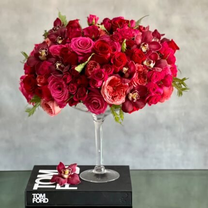 Elegant floral arrangement with red and pink roses, burgundy cymbidium orchids, and greens in a tall pedestal glass vase