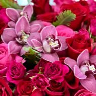 Elegant floral arrangement with red and pink roses, burgundy cymbidium orchids, and greens in a tall pedestal glass vase